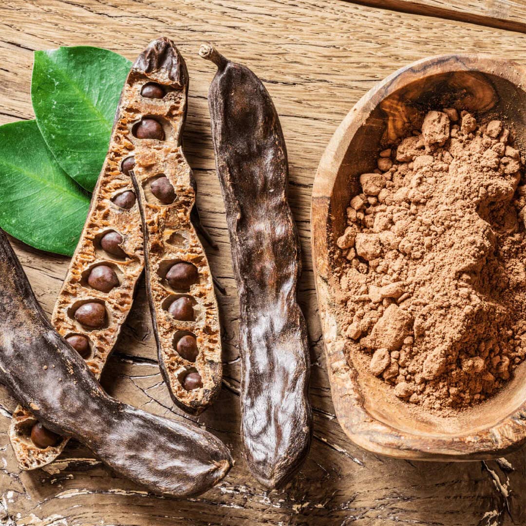 Baccelli di carruba, semi e polvere con foglie verdi su tavola di legno.